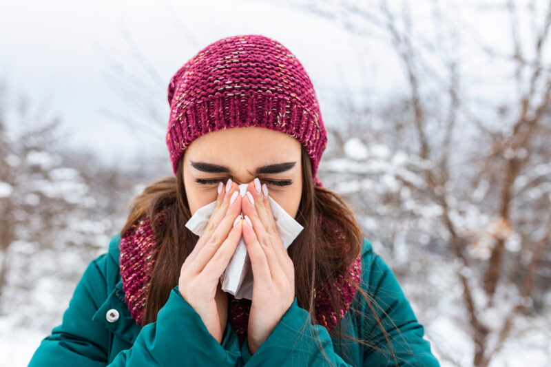 Young woman sick and sneeze into tissue paper. Girl blowing nose outdoors.Catching cold in winter. Upset gloomy woman with ill expression sneezes and has running nose