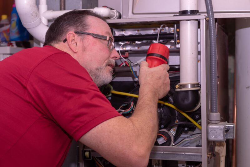 hvac technician leaning down to inspect the inside of a furnace with a flashlight