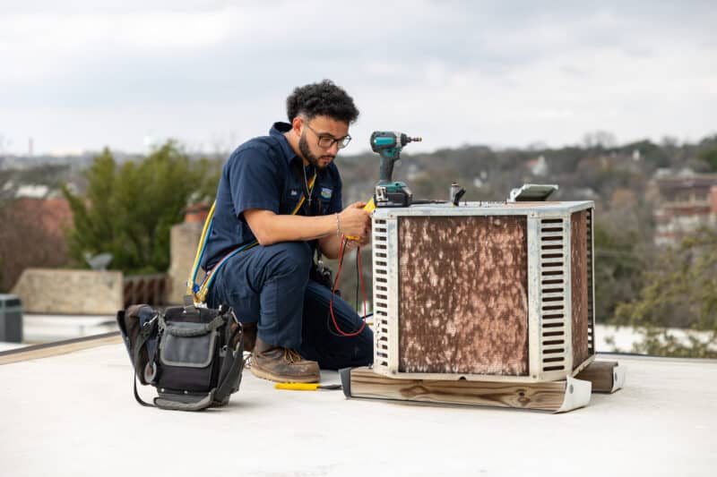 wright ac technician crouching down to perform repairs on a commercial outdoor hvac unit on top of a commercial roof, tools surrounding him to complete the job