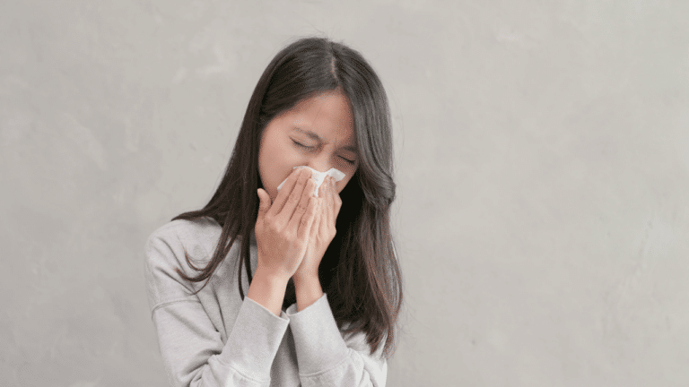 A woman with long dark hair sneezing into a tissue against a gray background.
