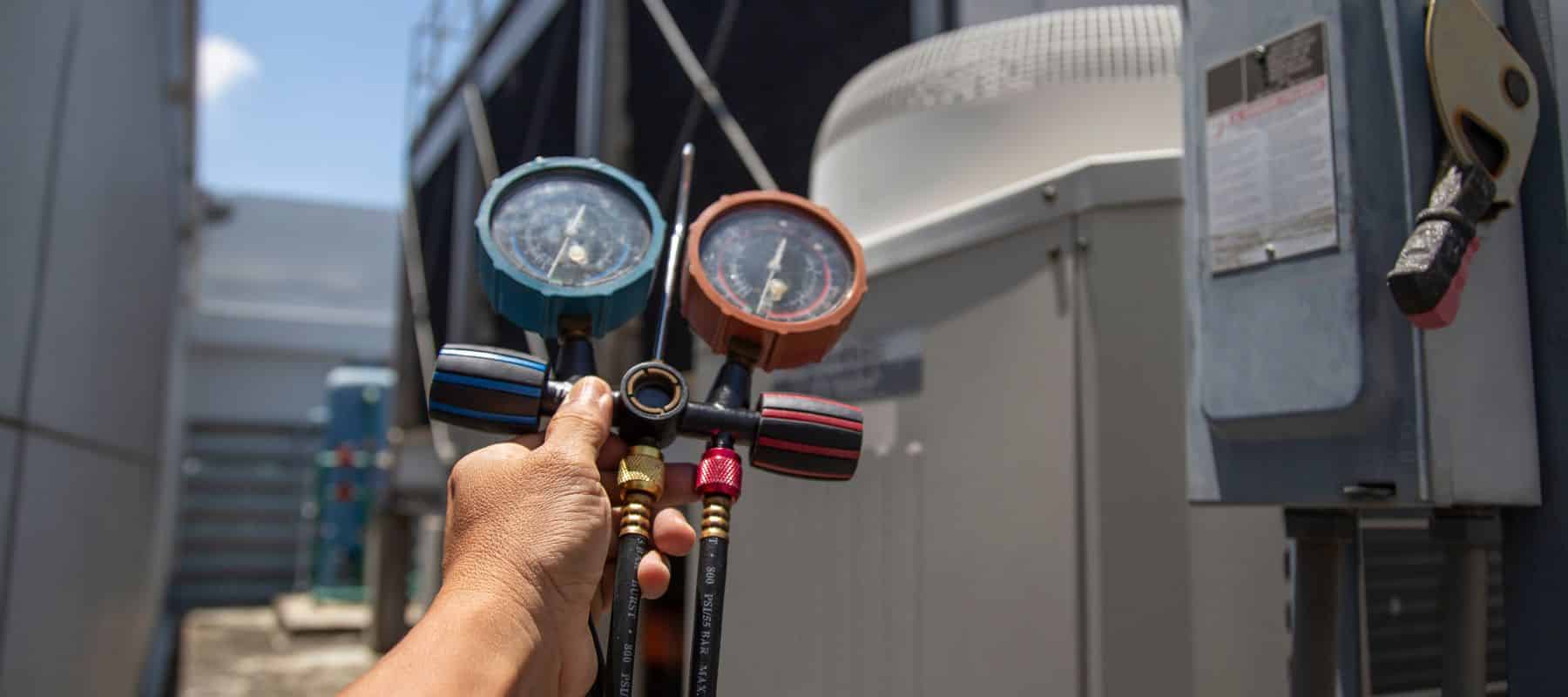 Close-up view of an HVAC technician's hand holding manifold gauges with pressure readings, used for checking air conditioning system performance, set against the backdrop of outdoor HVAC units