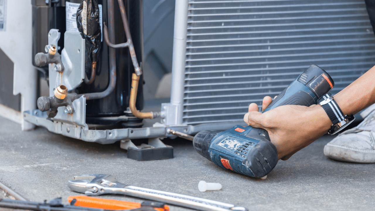 A man holding a power drill performing repairs on an AC unit.