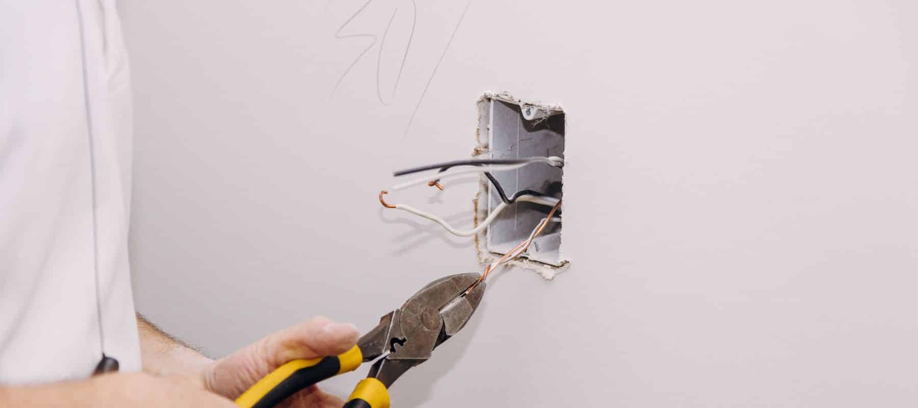 Electrician uses pliers to work on exposed wiring inside a wall outlet box