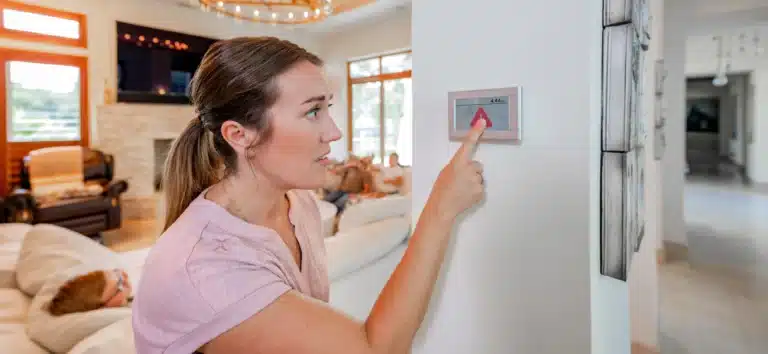 A woman adjusts the home thermostat while her family relaxes in the background