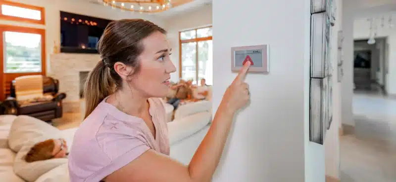 A woman adjusts the home thermostat while her family relaxes in the background