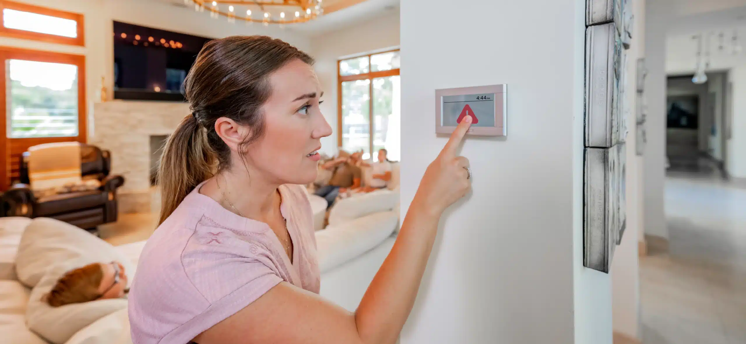 A woman adjusts the home thermostat while her family relaxes in the background