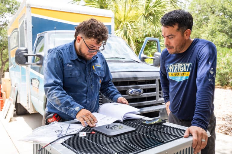 Two Wright Home Services HVAC technicians reading a manual over an outdoor condenser with a truck in the background.
