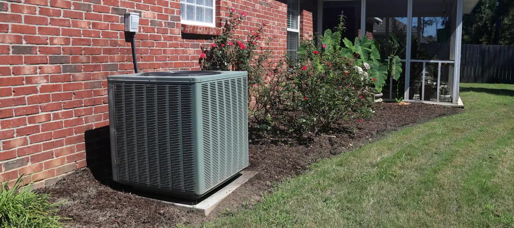 Outdoor air conditioning unit beside a red brick house surrounded by a garden with blooming bushes